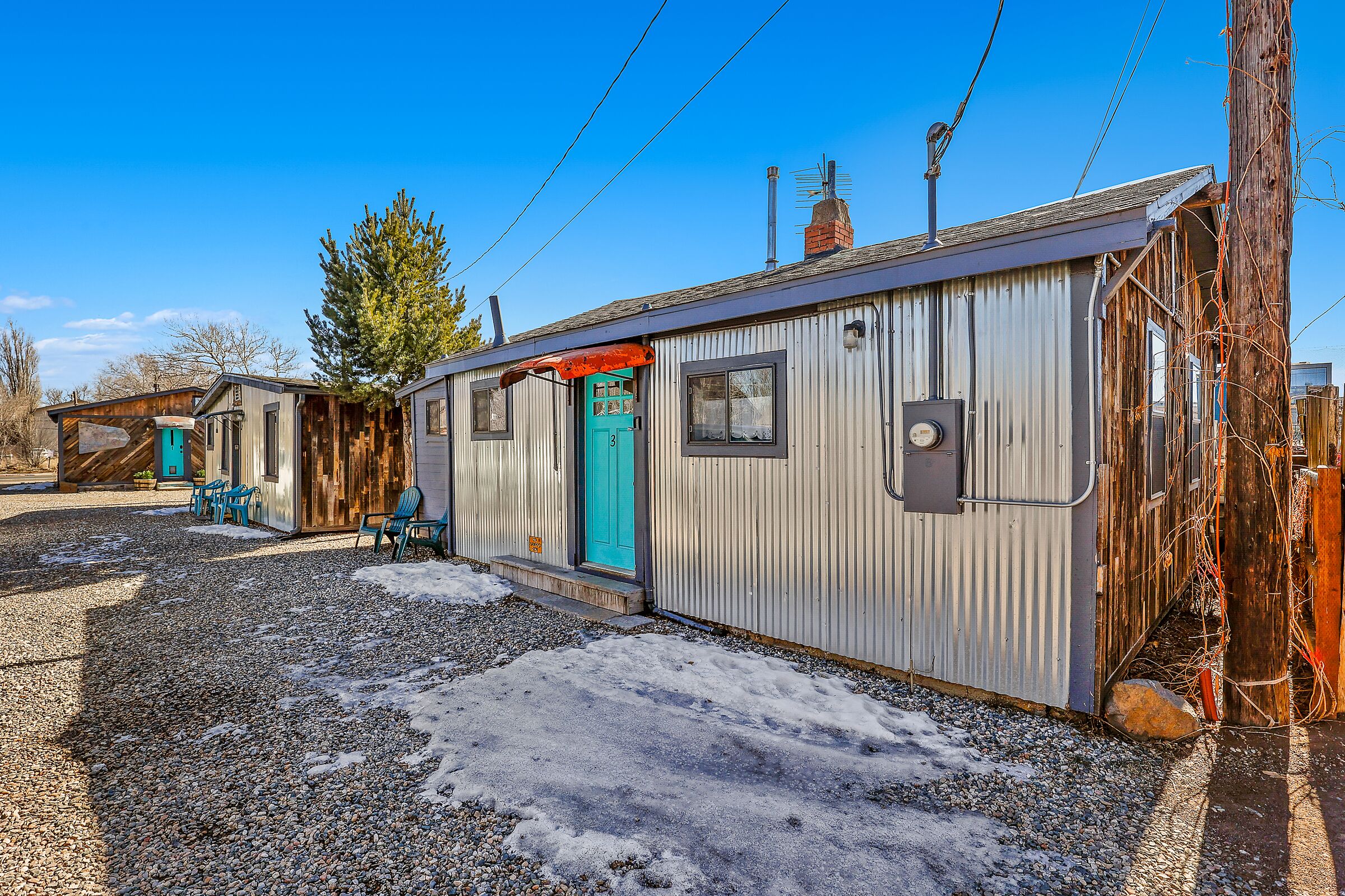 A small, single-story building with a metal exterior, a green door, and a gravel driveway.