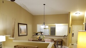 A living room with a sofa, a coffee table, and a dining table. There is a kitchen in the background with cabinets and a microwave.