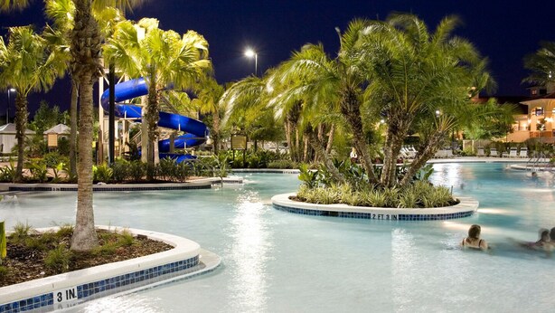 A nighttime swimming pool area with palm trees and a blue water slide.