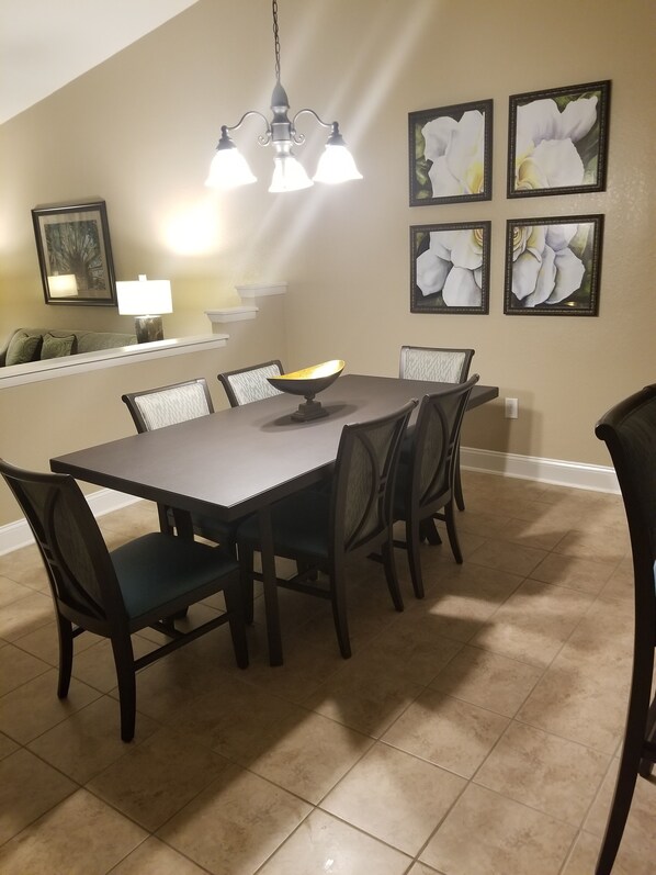 Un comedor con una mesa rectangular, seis sillas, una lámpara de araña y cuadros con motivos florales en la pared.