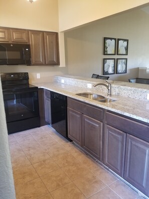 A kitchen with dark wooden cabinets, a granite countertop, a stainless steel sink, and a black dishwasher.