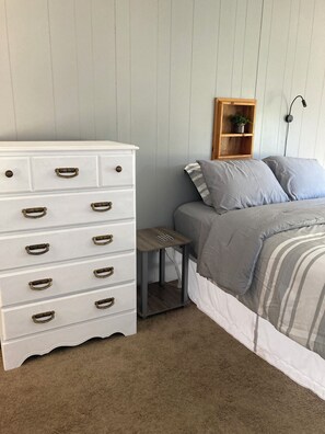A bedroom with a white dresser with multiple drawers and brass handles, a small wooden nightstand, a bed with grey bedding, and a wooden shelf with a plant.