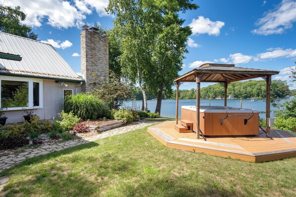 A lakeside property with a hot tub, wooden deck, stone pathway, and a gazebo.