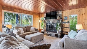 A cozy living room with a stone fireplace, wooden ceiling, and large windows overlooking a garden.