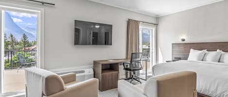 A hotel room with a bed, two armchairs, a desk with a chair, a flat-screen TV, and a large window with a mountain view.