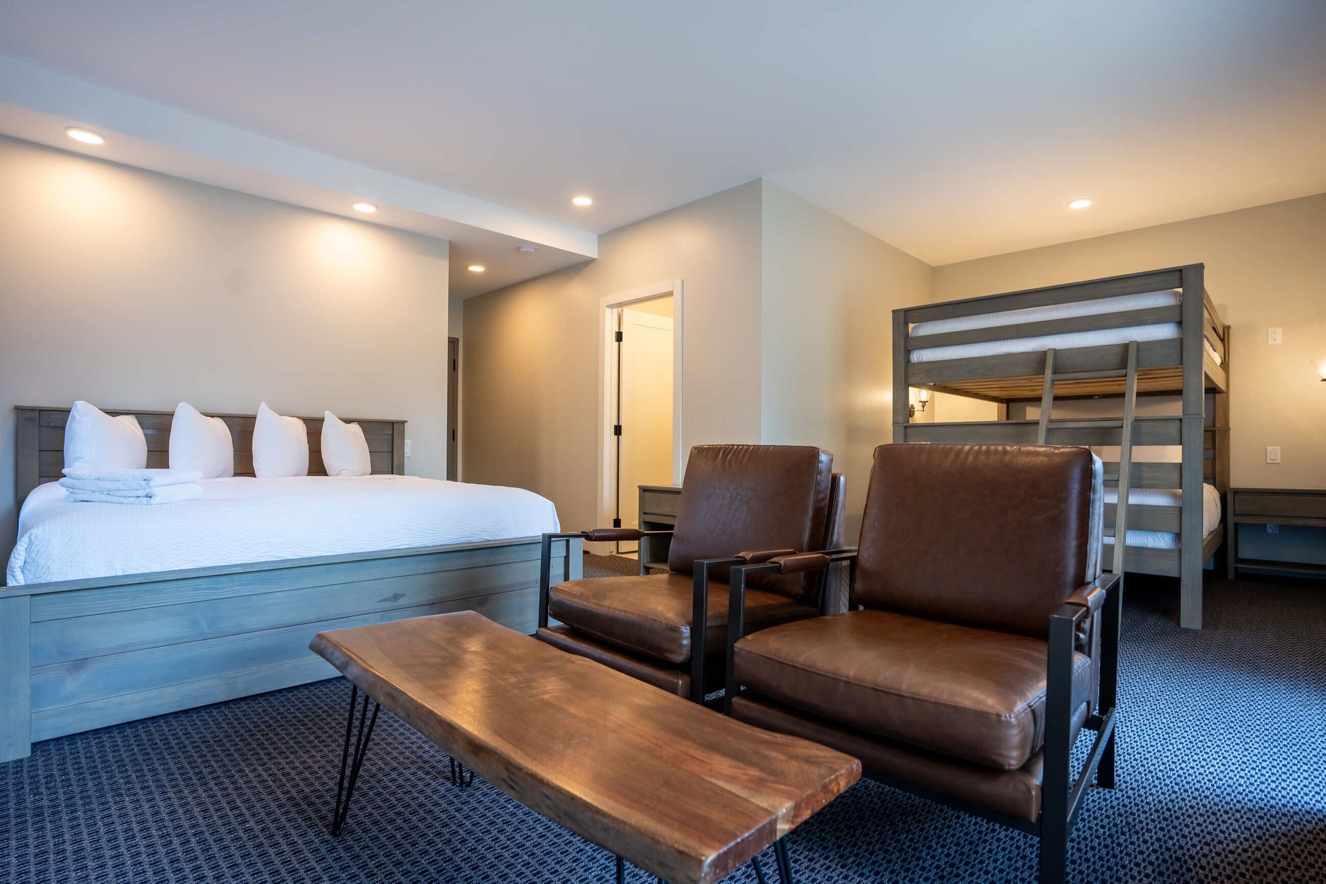 Una habitación de hotel con una cama, dos sillones de cuero marrón, un banco de madera y una cama individual al fondo.