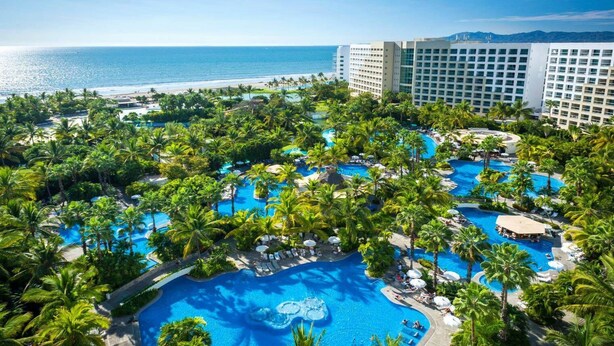 A resort with multiple swimming pools surrounded by palm trees, a beach, and high-rise buildings in the background.