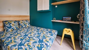 A bedroom with a teal wall, a wooden headboard, a bed with a floral bedspread, a yellow stool, a shelf with a laptop, and a patterned curtain.
