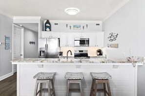 A kitchen with white cabinetry, a large island with stools, stainless steel appliances, and decorative wall art. - Regency Isle 307 | Beachfront | Pools, Hot Tubs and more! | My Beach Getaways (Orange Beach)