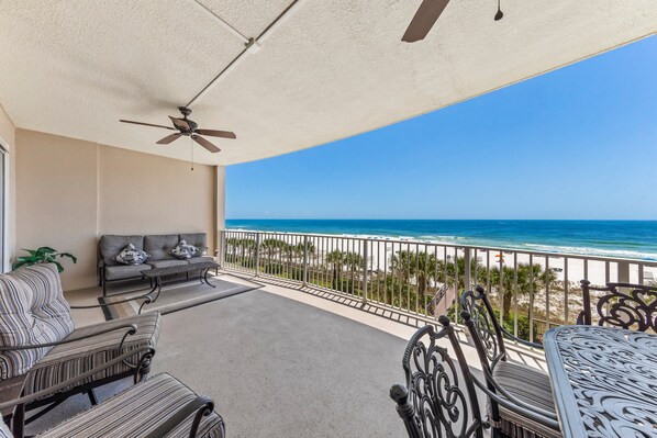 A balcony with a ceiling fan, a sofa, and a dining table with chairs. The balcony overlooks a beach with a railing.