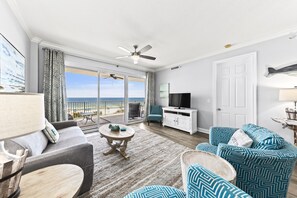 A spacious living room with a large window, a ceiling fan, a flat-screen TV, and a balcony with a view of the ocean. - Regency Isle 307 | Beachfront | Pools, Hot Tubs and more! | My Beach Getaways (Orange Beach)