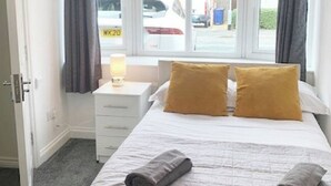 A hotel room with a bed, two pillows, a bedside lamp, a window with curtains, and a view of the street outside. - 5 BEDROOM / 4 BATHROOM MODERN DETACHED  wifi  Garden  Parking  Quiet location (Newcastle-under-Lyme)