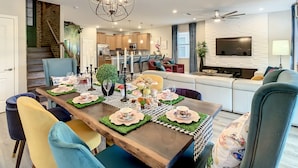 Une cuisine moderne équipée d’appareils électroménagers en acier inoxydable, un coin repas avec une table en bois pour quatre personnes, et un salon avec un canapé et un téléviseur.