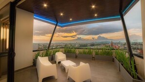 A modern rooftop terrace with white chairs and tables, a view of the cityscape, and a sunset. - Missum Versatile Living (San Andrés Cholula)