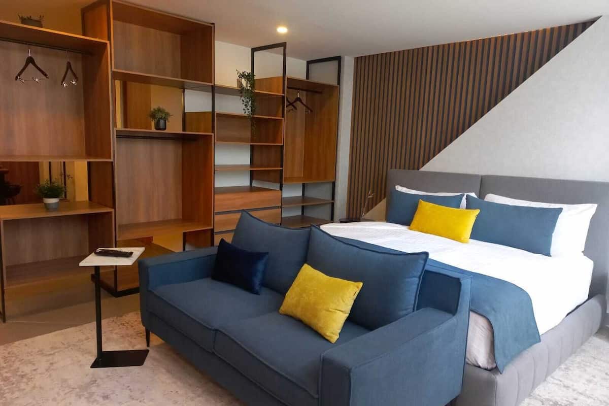 Un dormitorio moderno con una cama gris, almohadas azules y amarillas, un sofá azul y estanterías de madera.