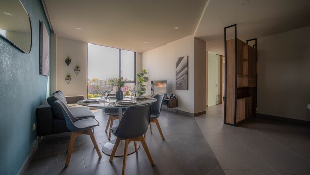 A modern dining area with a round table, chairs, and a view of the cityscape through large windows. - Missum Versatile Living (San Andrés Cholula)
