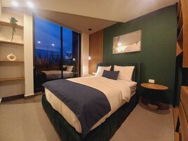 Un dormitorio moderno con una cama grande, una mesita de noche, una pequeña mesa redonda, un estante en la pared y un ventanal con vistas al cielo.