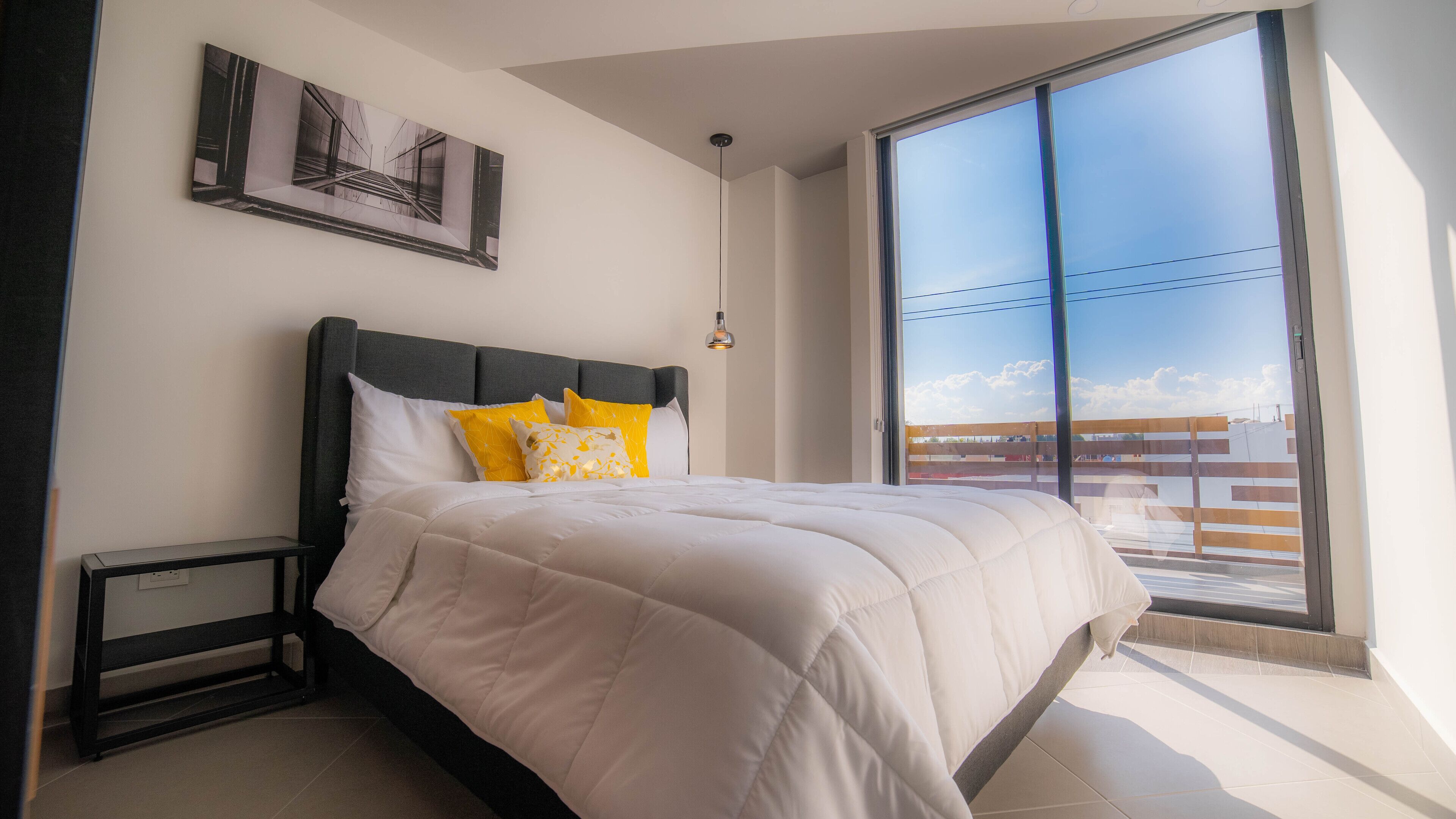 A bedroom with a large window, a bed with white bedding and yellow pillows, a bedside table, and a framed picture on the wall.