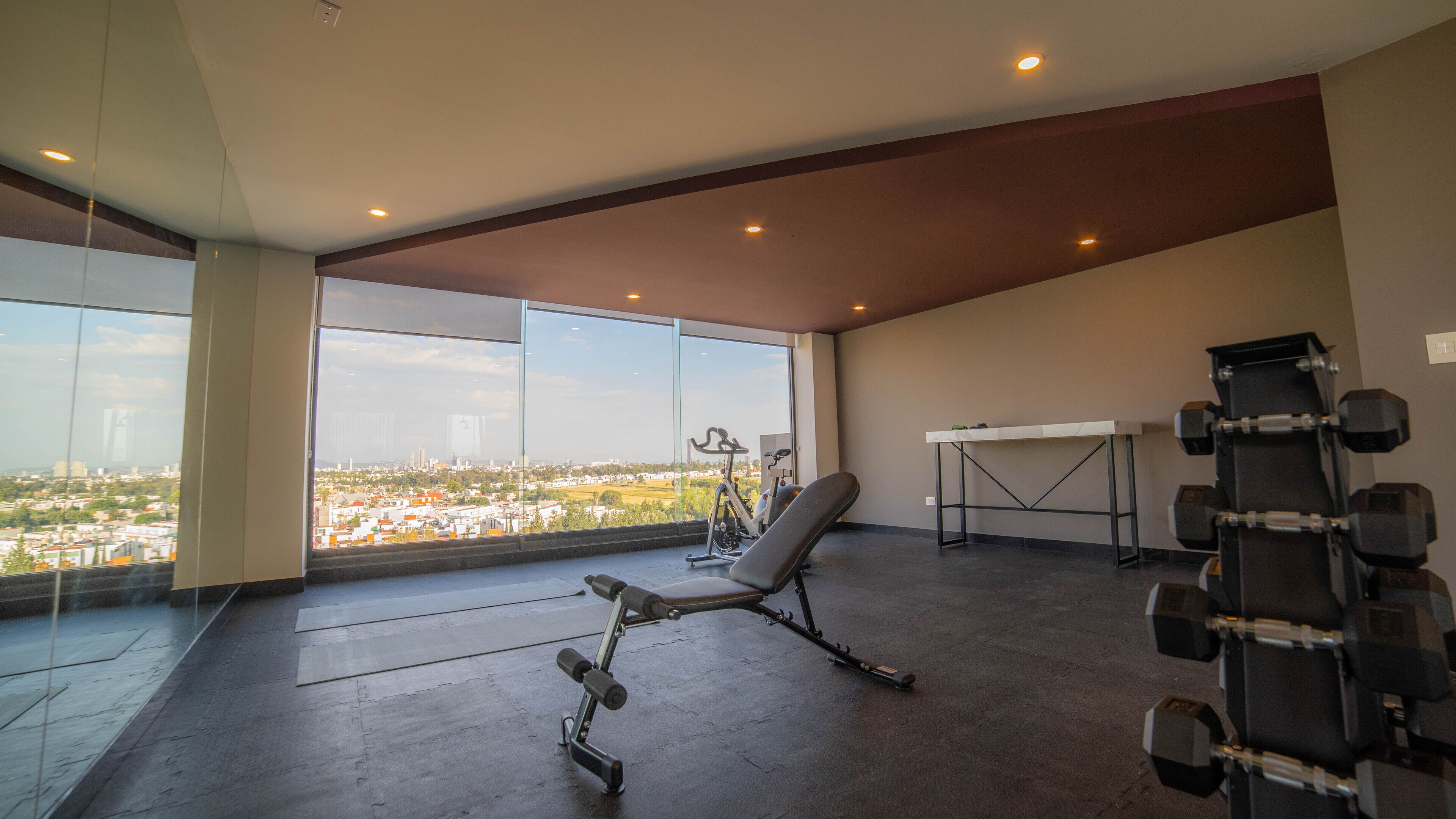 Amplia habitación con un gran ventanal que ofrece vistas a la ciudad, un banco, un escritorio y un juego de pesas.