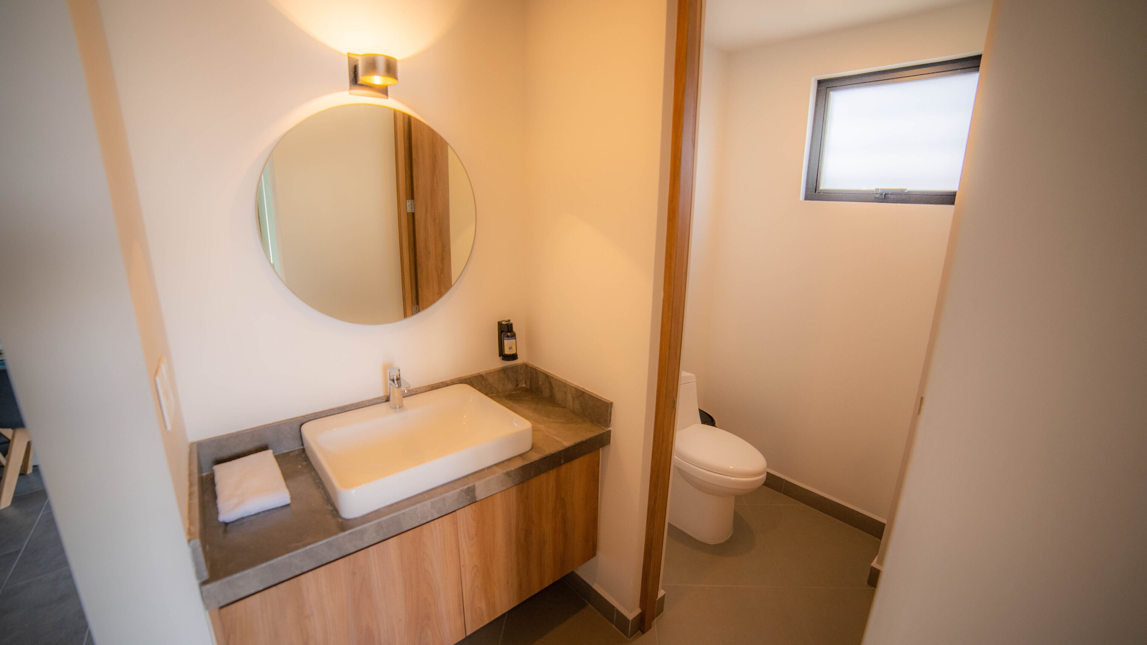 Ein Badezimmer mit einem weißen Waschbecken, einem Holzschrank, einem runden Spiegel mit Beleuchtung, einer Toilette und einem kleinen Fenster.