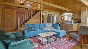 Ein gemütliches Wohnzimmer mit einem blauen Sofa, einem grünen Sessel und einem Holztisch. Eine Treppe führt zu einer oberen Ebene, und im Hintergrund ist eine Küche zu sehen.