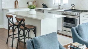 Uma cozinha moderna com armários brancos, uma ilha central, banquetas, fogão e uma janela grande.