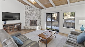 A modern living room with a stone fireplace, wooden ceiling beams, a flat-screen TV, and a large sliding glass door leading to an outdoor area. - Newly Rennovated and Air Conditioned Townhouse -1 mile from Skiing (Incline Village)
