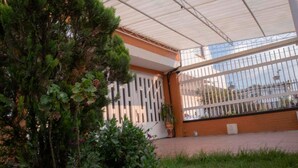 Un espace extérieur abrité, délimité par une clôture en métal, agrémenté d’un pot de fleurs et bordé d’un bâtiment à la façade blanche et aux ornements orange.
