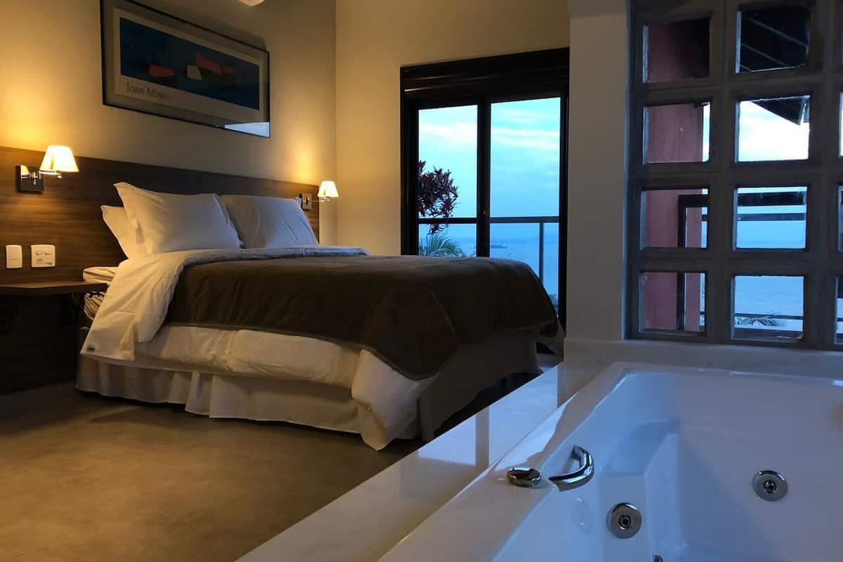 Quarto com cama grande, banheira e vista para o mar por uma janela ampla.