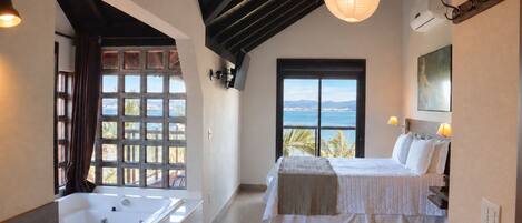 A bedroom with a large window, a bed, a bathtub, and a view of the sea.