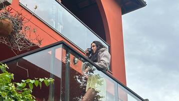 Uma pessoa em uma varanda com guarda-corpo de vidro, com vista para um edifício com fachada vermelha e uma estrutura em arco.