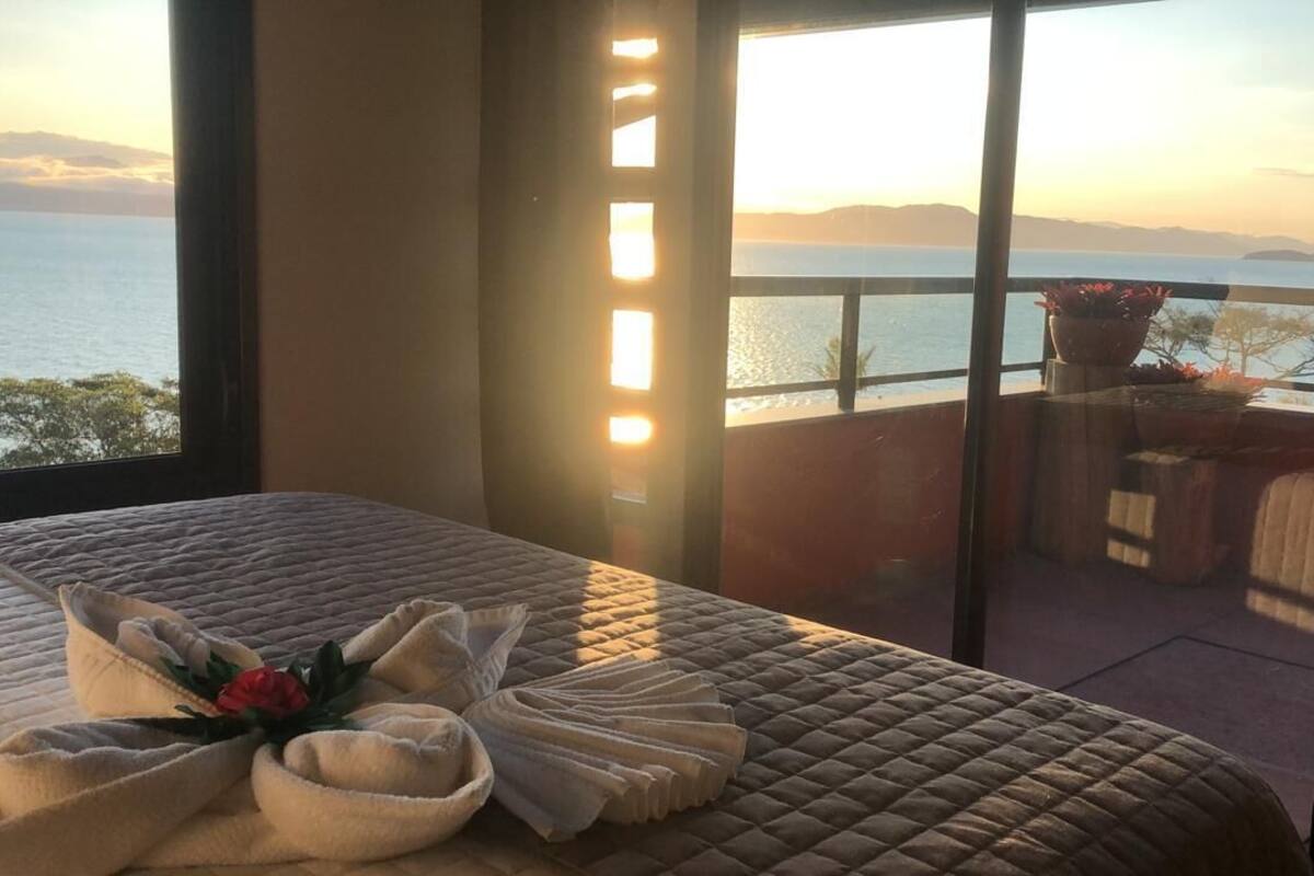 Quarto com cama grande, vista para o mar e varanda com assentos e plantas.