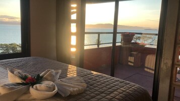 Un dormitorio con una cama grande, vista al mar y un balcón con mobiliario y plantas.