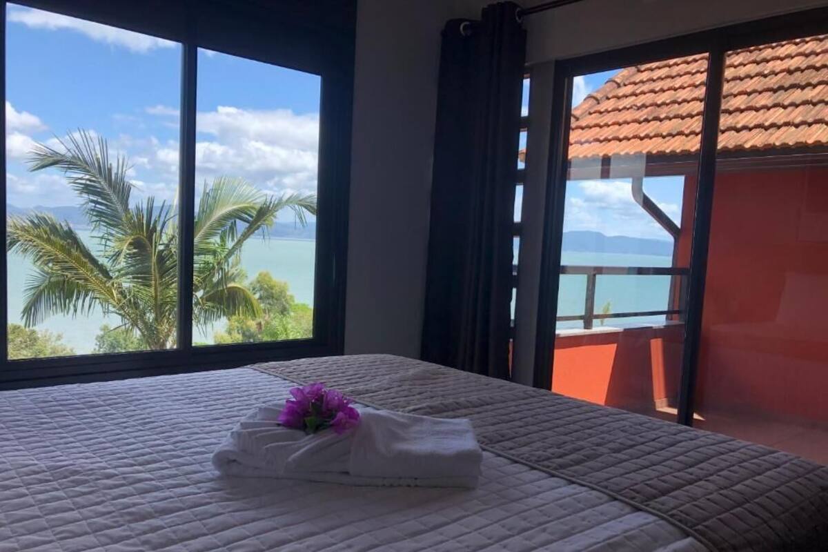 Cama com toalha dobrada e flor roxa, janela com vista para palmeiras e um corpo de água, e varanda com telhado vermelho.