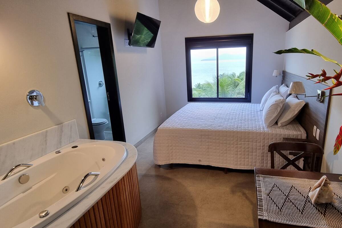 Quarto com uma janela grande, cama, mesa de cabeceira, cadeira, TV fixada na parede e banheiro com banheira.