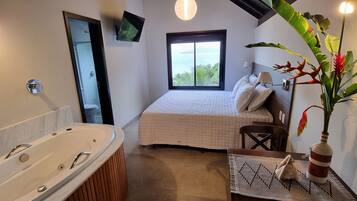 Un dormitorio con una ventana grande, una cama, una mesita de noche, una silla, un televisor montado en la pared y un baño con bañera.