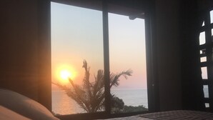 A bedroom with a large window overlooking a sunset over the sea, a bed with a quilted cover, and a palm tree visible outside. - Refúgio do Cacupé (Florianópolis)