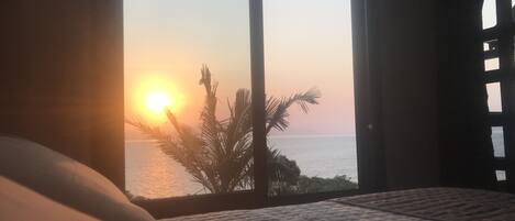 A bedroom with a large window overlooking a sunset over the sea, a bed with a quilted cover, and a palm tree visible outside.