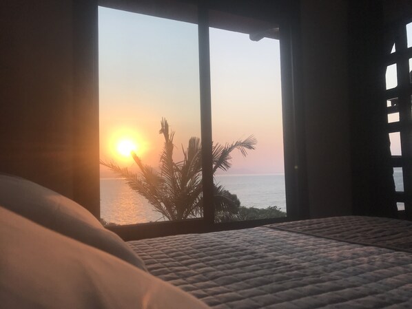 Un dormitorio con un amplio ventanal que da a una puesta de sol sobre el mar, una cama con cubrecama acolchado y una palmera visible desde el exterior.