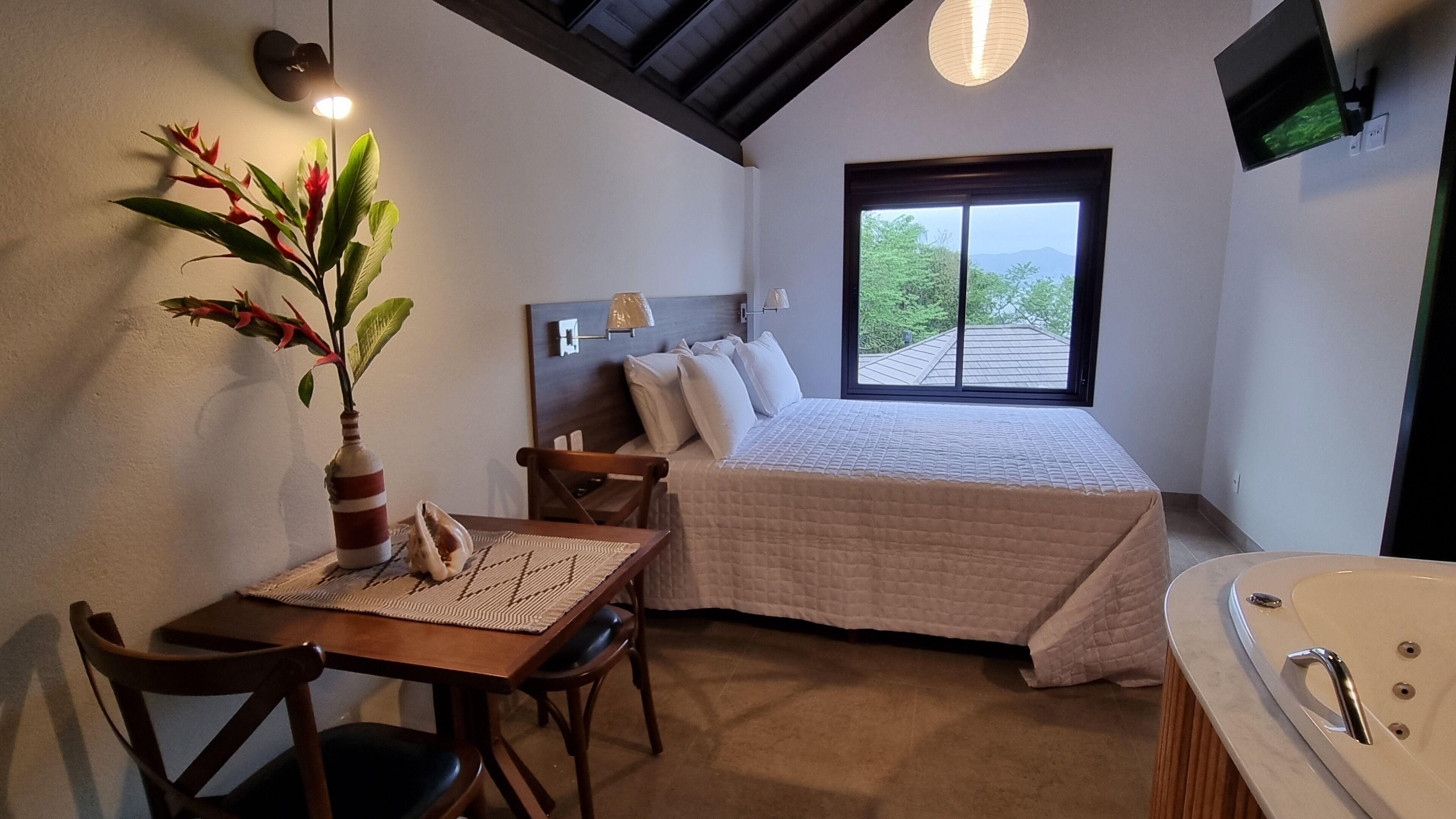 A bedroom with a bed, a desk, a chair, a plant, a TV, and a bathtub.