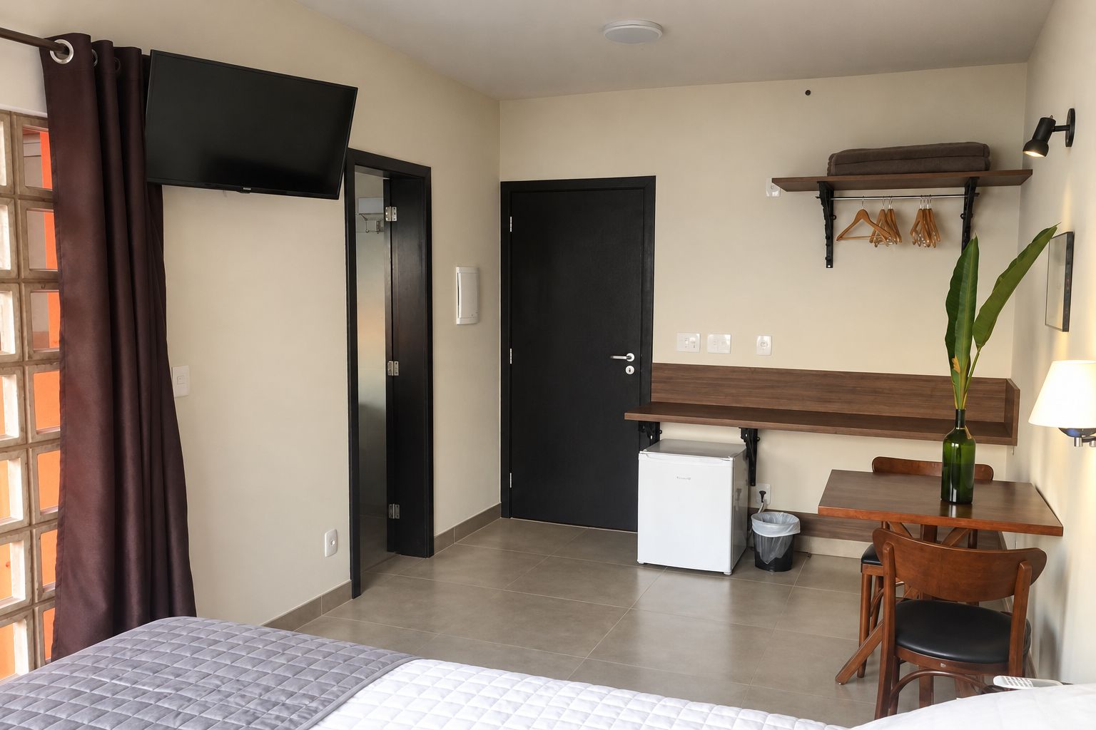 A hotel room with a bed, a desk, a chair, a small refrigerator, a trash bin, a plant, and a window with curtains.