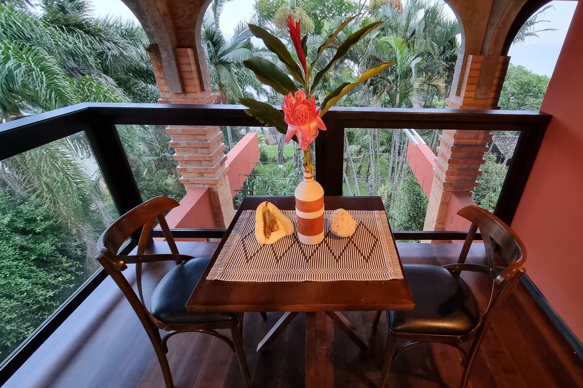 Uma mesa com um vaso de flores, um jogo americano estampado e duas cadeiras em uma varanda com vista para o verde.