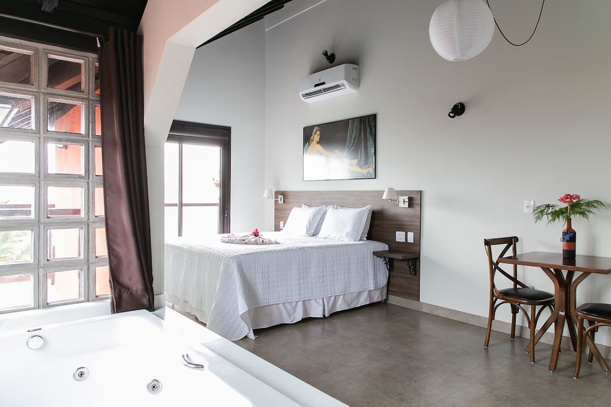 Quarto com uma cama grande, mesa de cabeceira, cadeira, pequena mesa e janela com cortinas.