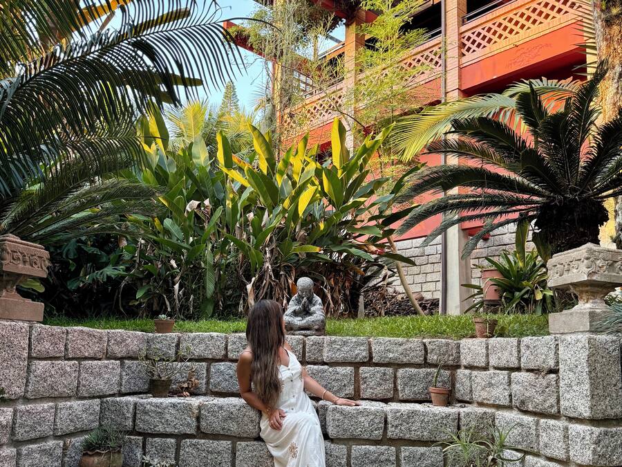 A person sitting on stone steps in a garden with tropical plants and a building with red and brown architecture in the background.