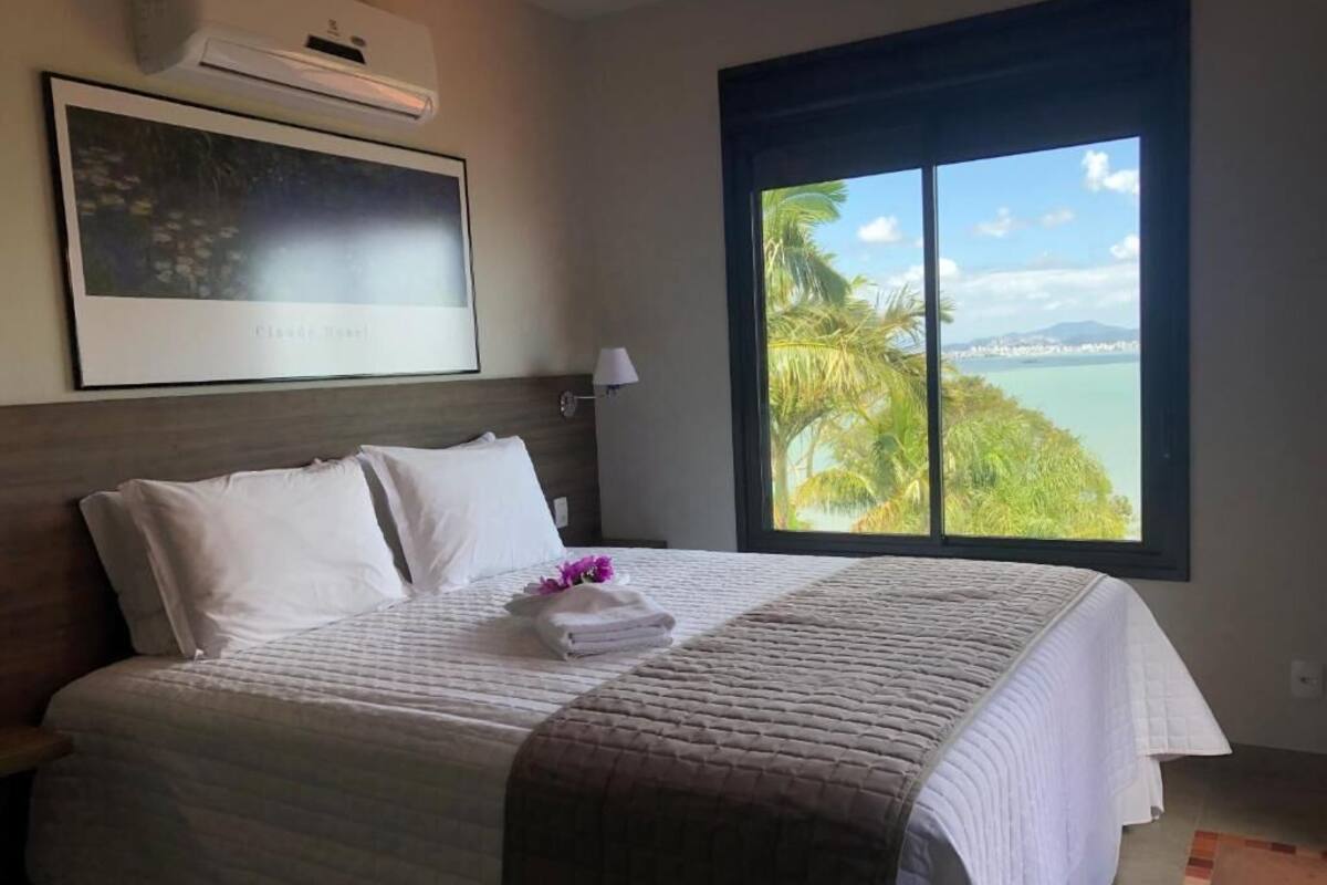 Quarto de hotel com cama, uma janela ampla com vista para palmeiras e água, e um quadro na parede.