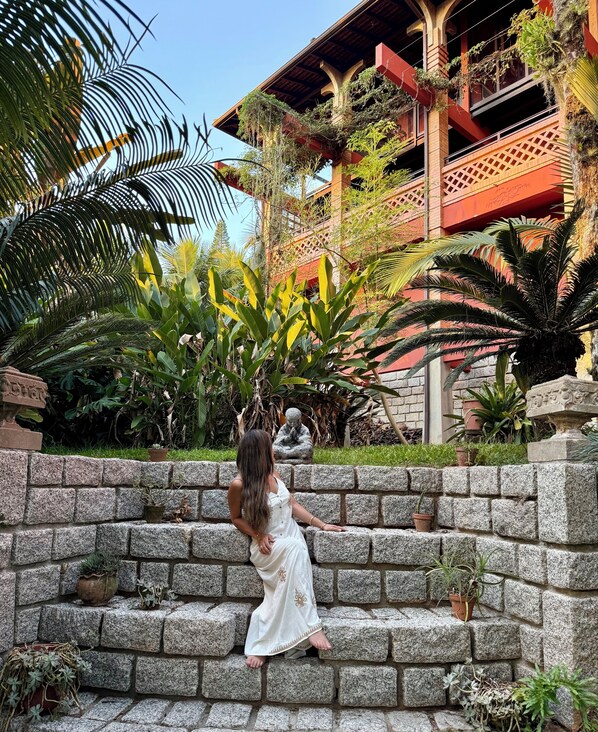 A woman in a white dress sitting on stone steps with potted plants and a building with red and brown architecture in the background. - Refúgio do Cacupé (Florianópolis)