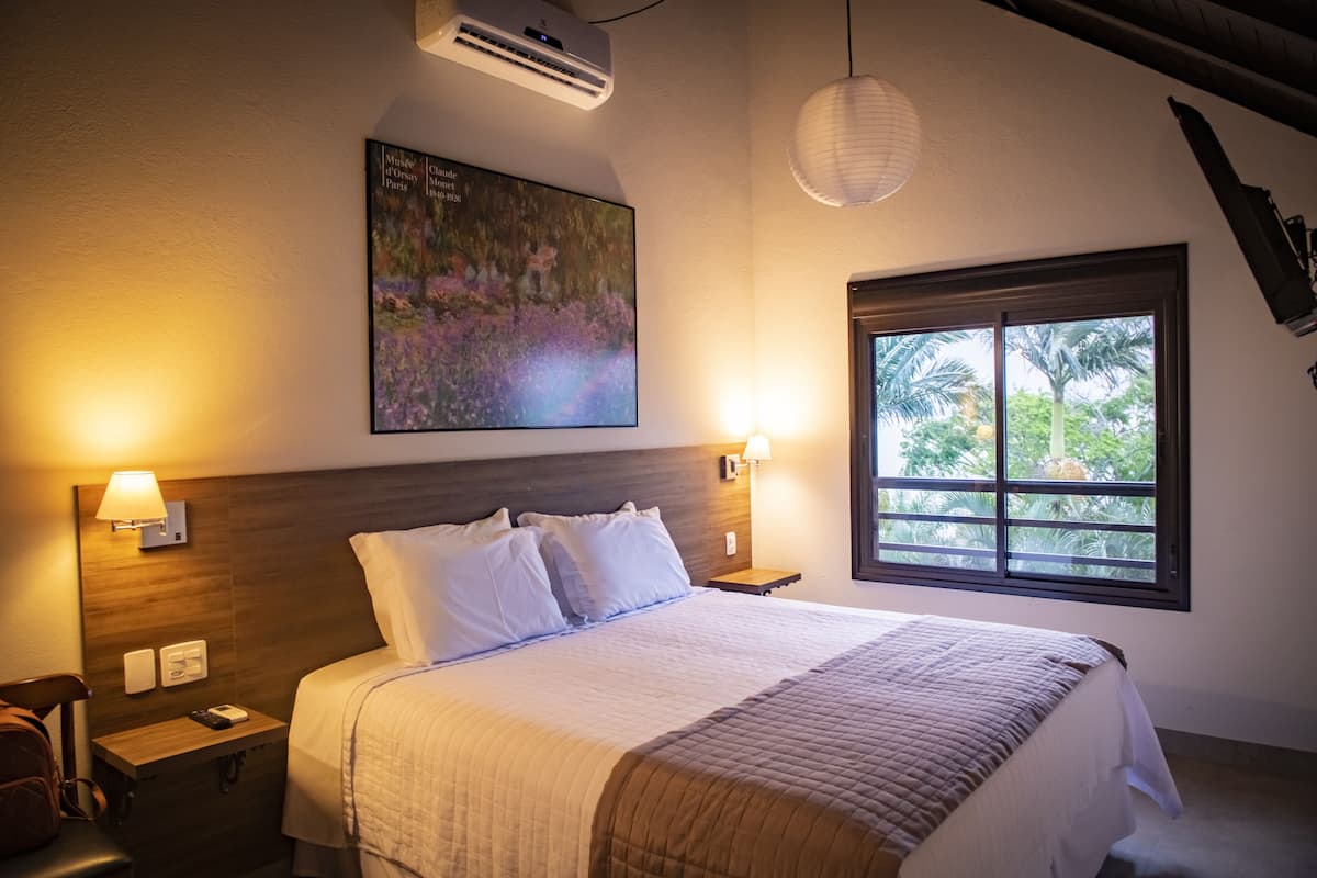 Quarto de hotel com cama, mesas de cabeceira, ar condicionado fixado na parede, janela com vista para palmeiras e um quadro na parede.