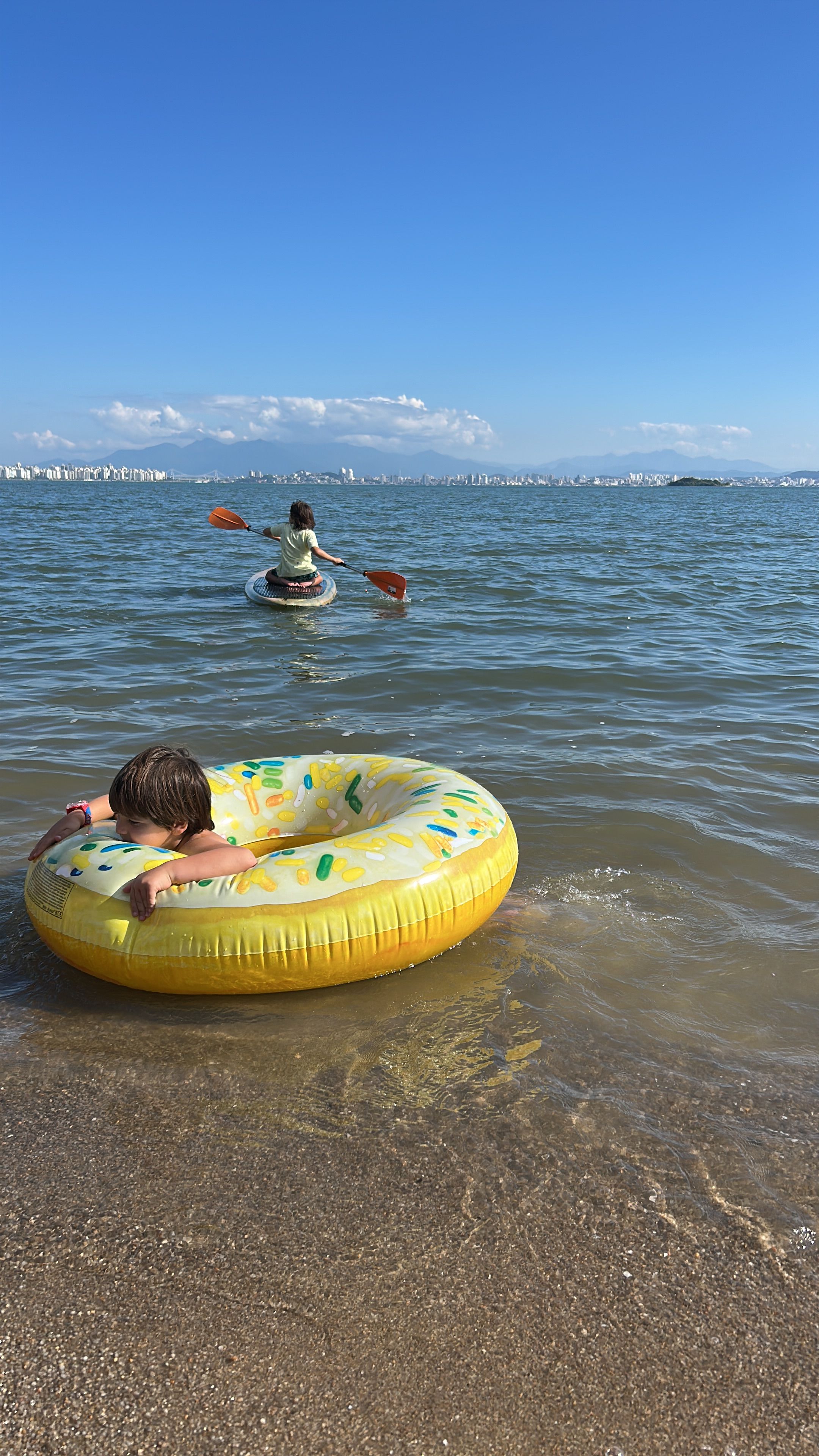 A person floating on a yellow inflatable ring in shallow water with a cityscape in the background.
