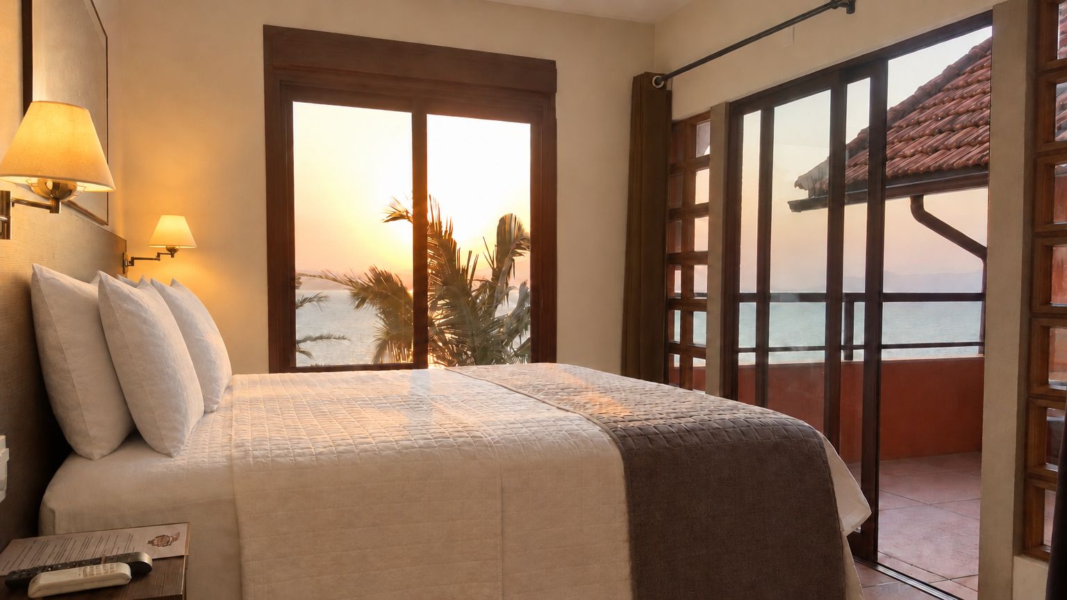 Una camera d'albergo con un letto, un comodino, una lampada, una finestra vista mare e una porta che dà su un balcone.