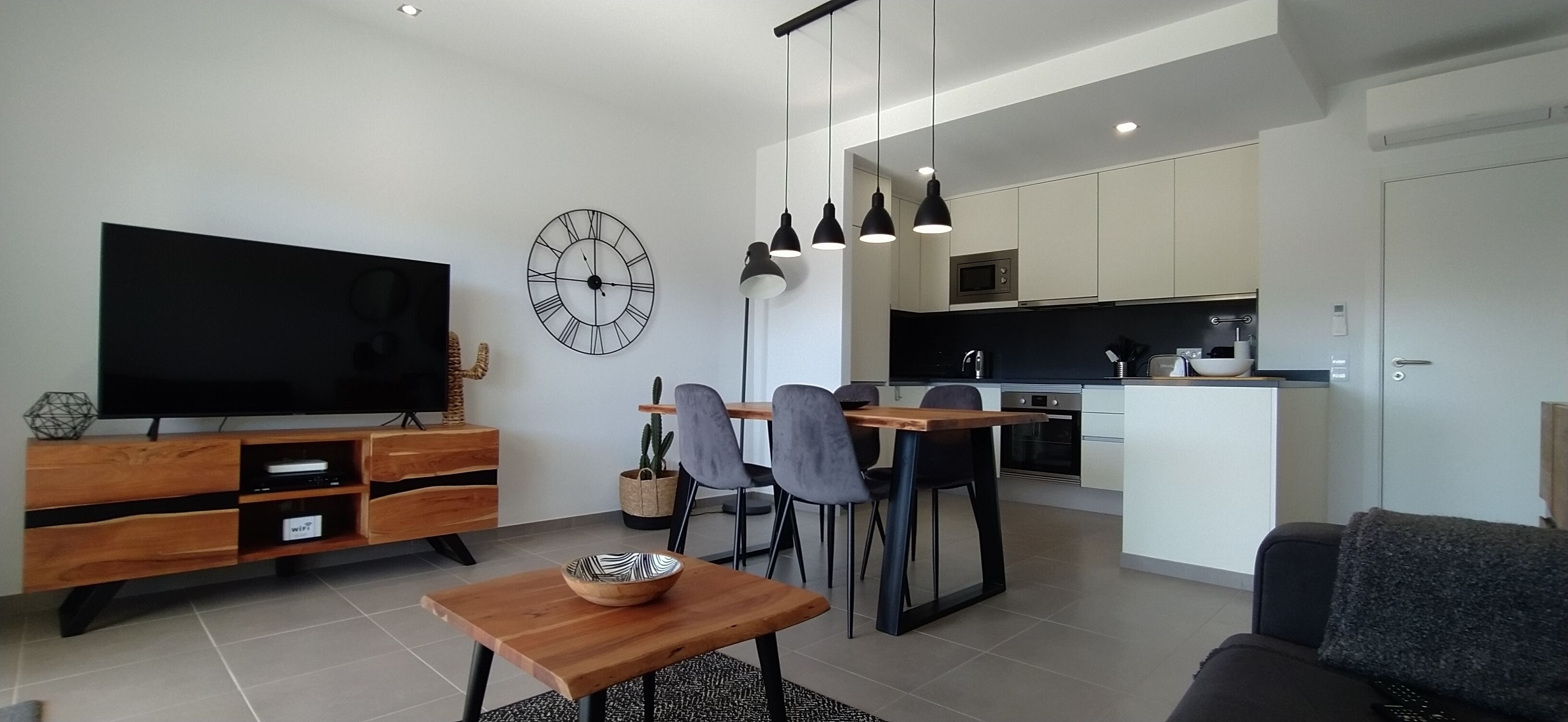 A modern living room with a flat-screen TV, wooden furniture, a dining area with chairs, and a kitchen with white cabinets and a black countertop.